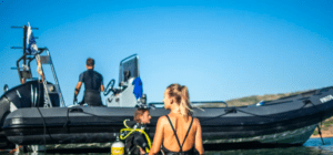 Un grupo se prepara con entusiasmo para una aventura de buceo cerca de su bote de goma en un día soleado, listos para experimentar el Bautismo de Buceo en Menorca en las aguas cristalinas. - ForTwo - Planes para parejas