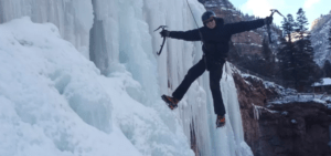 En Sierra Nevada, una persona escala una cascada congelada usando crampones y piolets. Las siluetas de los árboles la envuelven, capturando la emoción del "Bautismo de Escalada en Hielo en Sierra Nevada". - ForTwo - Planes para parejas