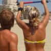 Dos personas en una playa, una levantando una barra colorida, con edificios y palmeras al fondo, es como si estuvieran comenzando su Bautismo de Kitesurf en Palma de Mallorca. - ForTwo - Planes para parejas