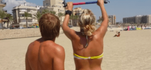 Dos personas en una playa, una levantando una barra colorida, con edificios y palmeras al fondo, es como si estuvieran comenzando su Bautismo de Kitesurf en Palma de Mallorca. - ForTwo - Planes para parejas