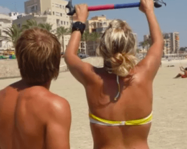 Dos personas en una playa, una levantando una barra colorida, con edificios y palmeras al fondo, es como si estuvieran comenzando su Bautismo de Kitesurf en Palma de Mallorca. - ForTwo - Planes para parejas