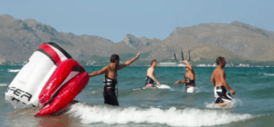 Cuatro hombres practican kitesurf en el océano con las escarpadas montañas de Palma de Mallorca como telón de fondo; uno de ellos domina las olas con maestría con una cometa roja y blanca. Disfruta de la experiencia con el Bautismo de Kitesurf en Palma de Mallorca. - ForTwo - Planes para parejas