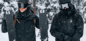 Dos esquiadores vestidos de negro sostienen sus esquís en medio de un bosque nevado de Sierra Nevada durante sus "Clases de Esquí en Sierra Nevada" mientras los copos de nieve caen suavemente a su alrededor. - ForTwo - Planes para parejas