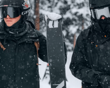 Dos esquiadores vestidos de negro sostienen sus esquís en medio de un bosque nevado de Sierra Nevada durante sus "Clases de Esquí en Sierra Nevada" mientras los copos de nieve caen suavemente a su alrededor. - ForTwo - Planes para parejas