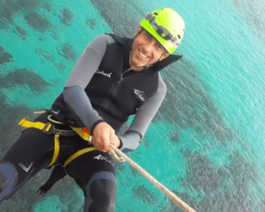 Una persona vestida con traje de neopreno y casco muestra una brillante sonrisa mientras disfruta del Coasteering en Manacor sobre sus cristalinas aguas azules. - ForTwo - Planes para parejas