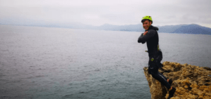 Con casco y traje de neopreno, una persona sonríe a la cámara desde un acantilado rocoso sobre el océano, emocionada por vivir una emocionante experiencia de Coasteering en Manacor. - ForTwo - Planes para parejas