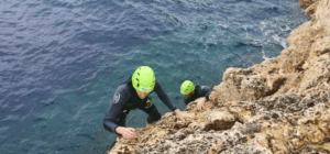 Dos aventureros con trajes de neopreno y cascos verdes escalan acantilados rocosos sobre aguas azules cristalinas, disfrutando de la emoción del Coasteering en Manacor a lo largo de su accidentada costa. - ForTwo - Planes para parejas