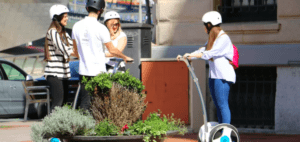 Cuatro conductores con casco se deslizan con soltura en Segways por una acera soleada cerca de una jardinera, capturando la vibrante esencia de Bilbao. Experimenta la alegría de los patinetes autoequilibrados con "Descubre Bilbao en Segway" en este pintoresco escenario. - ForTwo - Planes para parejas