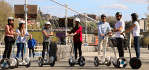Un grupo de ocho personas explora la naturaleza en segways con casco, disfrutando de vibrantes paisajes y un puente panorámico de fondo. Su aventura ofrece una forma única de descubrir el encanto de Bilbao con "Descubre Bilbao en Segway". - ForTwo - Planes para parejas
