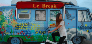 Una mujer en bicicleta frente a una furgoneta llena de grafitis con el cartel de Le Break resume el encanto ecléctico de Córdoba. Descubre la ciudad como nunca antes: vívela con Descubre Córdoba en Bici Eléctrica y vive cada momento de color. - ForTwo - Planes para parejas