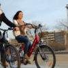 Dos personas recorren un sendero pavimentado con Descubre Córdoba en Bici Eléctrica, disfrutando de los encantadores edificios y la frondosa arboleda de fondo. Es un día ideal para descubrir nuevos lugares juntos. - ForTwo - Planes para parejas