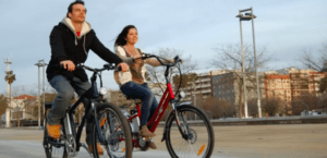 Dos personas recorren un sendero pavimentado con Descubre Córdoba en Bici Eléctrica, disfrutando de los encantadores edificios y la frondosa arboleda de fondo. Es un día ideal para descubrir nuevos lugares juntos. - ForTwo - Planes para parejas