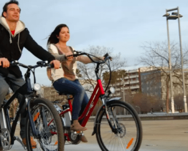 Dos personas recorren un sendero pavimentado con Descubre Córdoba en Bici Eléctrica, disfrutando de los encantadores edificios y la frondosa arboleda de fondo. Es un día ideal para descubrir nuevos lugares juntos. - ForTwo - Planes para parejas