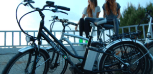Dos mujeres se sientan en una cornisa en Córdoba, charlando y riendo detrás de bicicletas eléctricas Descubre Córdoba en Bici aparcadas, su alegría mezclándose con el bullicio de la ciudad. - ForTwo - Planes para parejas