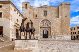 Explora Mahón y su rica historia con "Descubre Mahón a Tu Ritmo", donde podrás admirar la histórica fachada de la iglesia de piedra y la majestuosa estatua de tres caballos en primer plano, todo bajo un cielo parcialmente nublado, ideal para turistas que aprecian la belleza arquitectónica. - ForTwo - Planes para parejas