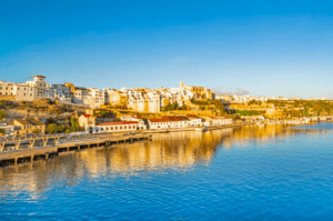 Experimente el pintoresco Mahón con "Descubre Mahón a Tu Ritmo", donde los edificios blancos se funden con un sereno mar azul bajo un cielo despejado, ideal para el turismo. - ForTwo - Planes para parejas