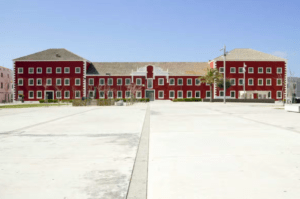 Explora la belleza de Mahón con "Descubre Mahón a Tu Ritmo", que presenta un gran edificio rojo y blanco con una fachada simétrica, ubicado en una plaza vacía bajo un cielo azul claro, ideal para el turismo tranquilo. - ForTwo - Planes para parejas
