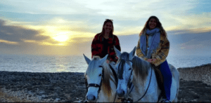 Dos personas pasean a caballo por la rocosa costa de Menorca al atardecer, bajo un cielo nublado. Experimenta la belleza de este paisaje costero con Descubre Menorca a Caballo. - ForTwo - Planes para parejas