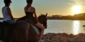 Dos personas cabalgando cerca de una costa rocosa de Menorca, disfrutando de la puesta de sol sobre el agua, mientras experimentan la impresionante belleza de la naturaleza con "Descubre Menorca a Caballo". - ForTwo - Planes para parejas