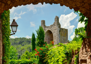 Sobre un antiguo arco de piedra, la exuberante vegetación de Girona y sus vibrantes flores rojas contrastan con un cielo azul, encarnando el encanto perdurable de Descubre Pueblos Medievales desde Girona. - ForTwo - Planes para parejas