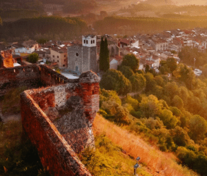 Vista aérea de Hostalric que muestra el Castell d'Hostalric, un encantador castillo de piedra, y su iglesia, rodeados de exuberante vegetación al atardecer. Descubre este pintoresco paisaje donde la historia se fusiona con la belleza natural con Descubre el Castell d'Hostalric. - ForTwo - Planes para parejas