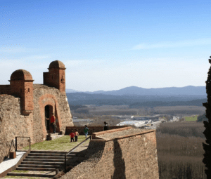 Un castillo medieval de piedra con puertas arqueadas, similar a Descubre el Castell d’Hostalric, domina las montañas y un valle bajo un cielo azul claro. - ForTwo - Planes para parejas