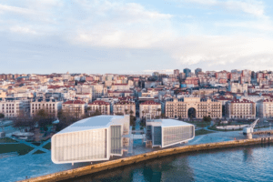 Una vista aérea presenta modernos edificios frente al mar con la elegancia de "Descubre el Centro Botín" enclavado en el paisaje urbano, todo bajo un cielo azul claro. - ForTwo - Planes para parejas