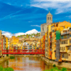 Edificios coloridos bordean el río Onyar en Girona, que aparece en "Descubre el corazón histórico de Girona", con un puente rojo y una iglesia al fondo. - ForTwo - Planes para parejas
