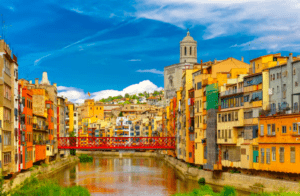 Edificios coloridos bordean el río Onyar en Girona, que aparece en "Descubre el corazón histórico de Girona", con un puente rojo y una iglesia al fondo. - ForTwo - Planes para parejas