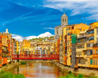 Edificios coloridos bordean el río Onyar en Girona, que aparece en "Descubre el corazón histórico de Girona", con un puente rojo y una iglesia al fondo. - ForTwo - Planes para parejas