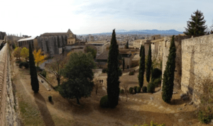 Experimente "Descubre el corazón histórico de Girona" con una vista panorámica de una antigua fortaleza, muros de piedra, árboles altos y el paisaje urbano al fondo: un verdadero corazón histórico. - ForTwo - Planes para parejas