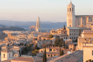 Un paisaje urbano iluminado por el sol de Girona, España, presenta edificios históricos y colinas distantes bajo un cielo despejado, como se captura en "Descubre el corazón histórico de Girona. - ForTwo - Planes para parejas