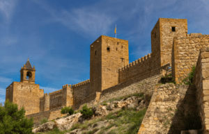Descubre la Historia y Naturaleza de Antequera" muestra una histórica fortaleza de piedra con torres y almenas bellamente recortadas contra un cielo azul claro. - ForTwo - Planes para parejas
