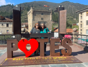 Dos personas posan detrás de un gran cartel con un corazón que dice "Descubre los Picos de Europa y Potes", frente al impresionante telón de fondo de las majestuosas colinas y los pintorescos edificios de los Picos de Europa. - ForTwo - Planes para parejas