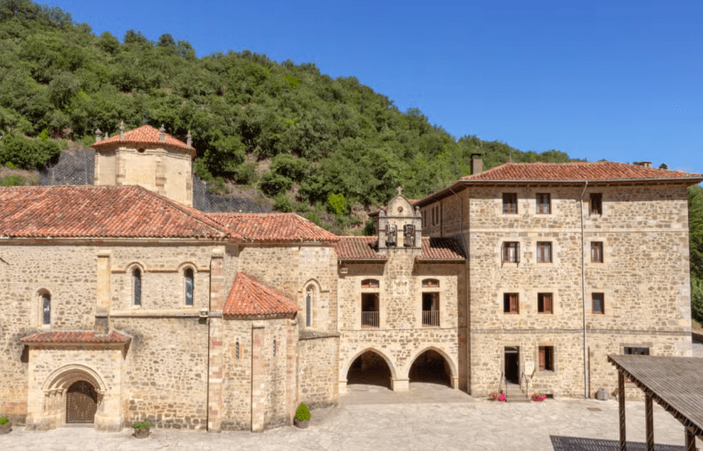 En el corazón de Potes, históricos edificios de piedra con tejados de tejas rojas se alzan entre la exuberante vegetación y los majestuosos picos de los Picos de Europa bajo un cielo azul despejado. Vívelo con "Descubre los Picos de Europa y Potes". - ForTwo - Planes para parejas