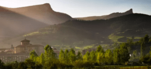 La tranquila escena del pueblo bajo un cielo soleado y despejado con montañas, árboles y una iglesia notable evoca la tranquilidad de una "Escapada al Salto del Nervión". - ForTwo - Planes para parejas