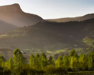 La tranquila escena del pueblo bajo un cielo soleado y despejado con montañas, árboles y una iglesia notable evoca la tranquilidad de una "Escapada al Salto del Nervión". - ForTwo - Planes para parejas