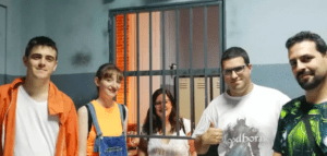 Cinco personas posan con una celda de prisión como telón de fondo en Escape Room Málaga: Escapad de una prisión encantada; uno está dentro, sonriendo como si acabara de descifrar el código definitivo. - ForTwo - Planes para parejas