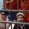 Dos individuos, uno con gorra de policía y el otro con gorra de prisionero, están tras las rejas, con dos más de pie detrás. Es como entrar en Escape Room Málaga: Escapada de una prisión encantada. - ForTwo - Planes para parejas