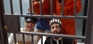 Dos individuos, uno con gorra de policía y el otro con gorra de prisionero, están tras las rejas, con dos más de pie detrás. Es como entrar en Escape Room Málaga: Escapada de una prisión encantada. - ForTwo - Planes para parejas