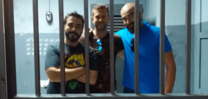 Tres hombres sonriendo tras las rejas de una celda, vestidos con ropa informal y con marcas de conteo en la pared, una escena que recuerda a Escape Room Málaga: Escapad de una prisión encantada. - ForTwo - Planes para parejas
