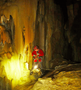 Vestido con equipo rojo de espeleología, una persona explora una cueva poco iluminada con estalactitas y el equipo esencial, encarnando la esencia de la Espeleología en la Sierra de las Nieves. - ForTwo - Planes para parejas