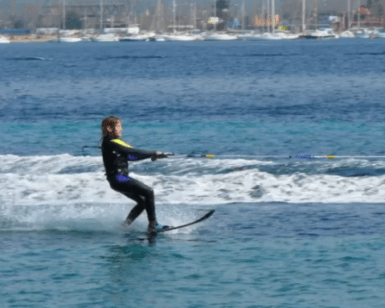 Una persona disfruta de su tiempo en Esquí Acuático en Pollença, deslizándose por el vibrante mar azul, mientras un encantador paisaje urbano y una flota de barcos crean un pintoresco telón de fondo. - ForTwo - Planes para parejas