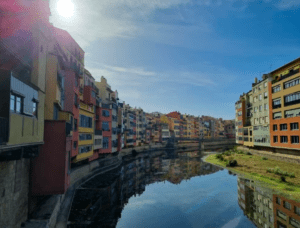 El sol brillante y el cielo azul iluminan los coloridos edificios junto al río, haciéndose eco de la vitalidad de la Excursión a Gerona y Figueres. - ForTwo - Planes para parejas