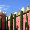 En Figueres, un edificio rojo presenta grandes esculturas de huevos en el techo, rodeado de altos cipreses y contra un cielo azul, ideal para una "Excursión a Gerona y Figueres". - ForTwo - Planes para parejas