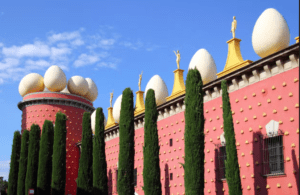 En Figueres, un edificio rojo presenta grandes esculturas de huevos en el techo, rodeado de altos cipreses y contra un cielo azul, ideal para una "Excursión a Gerona y Figueres". - ForTwo - Planes para parejas