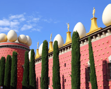 En Figueres, un edificio rojo presenta grandes esculturas de huevos en el techo, rodeado de altos cipreses y contra un cielo azul, ideal para una "Excursión a Gerona y Figueres". - ForTwo - Planes para parejas