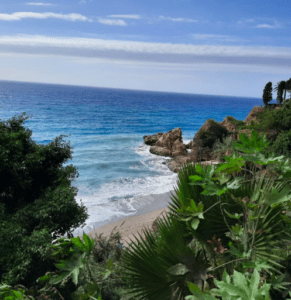 Disfrute de una vista impresionante de la costa rocosa cerca de Frigiliana durante la "Excursión a Nerja, Frigiliana y El Acebuchal", donde la exuberante vegetación domina el océano bajo un cielo parcialmente nublado. - ForTwo - Planes para parejas
