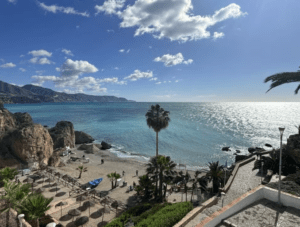 Disfrute de las vistas costeras con playas de arena, mar azul y acantilados rocosos cerca de Nerja en la "Excursión a Nerja, Frigiliana y El Acebuchal", donde un cielo parcialmente nublado realza la escena con palmeras y pasarelas que añaden encanto a su experiencia. - ForTwo - Planes para parejas