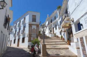 La estrecha calle, que evoca la excursión a Nerja, Frigiliana y El Acebuchal, está bordeada de edificios blancos y escalones de piedra, con una motocicleta enclavada bajo el cielo azul claro. - ForTwo - Planes para parejas